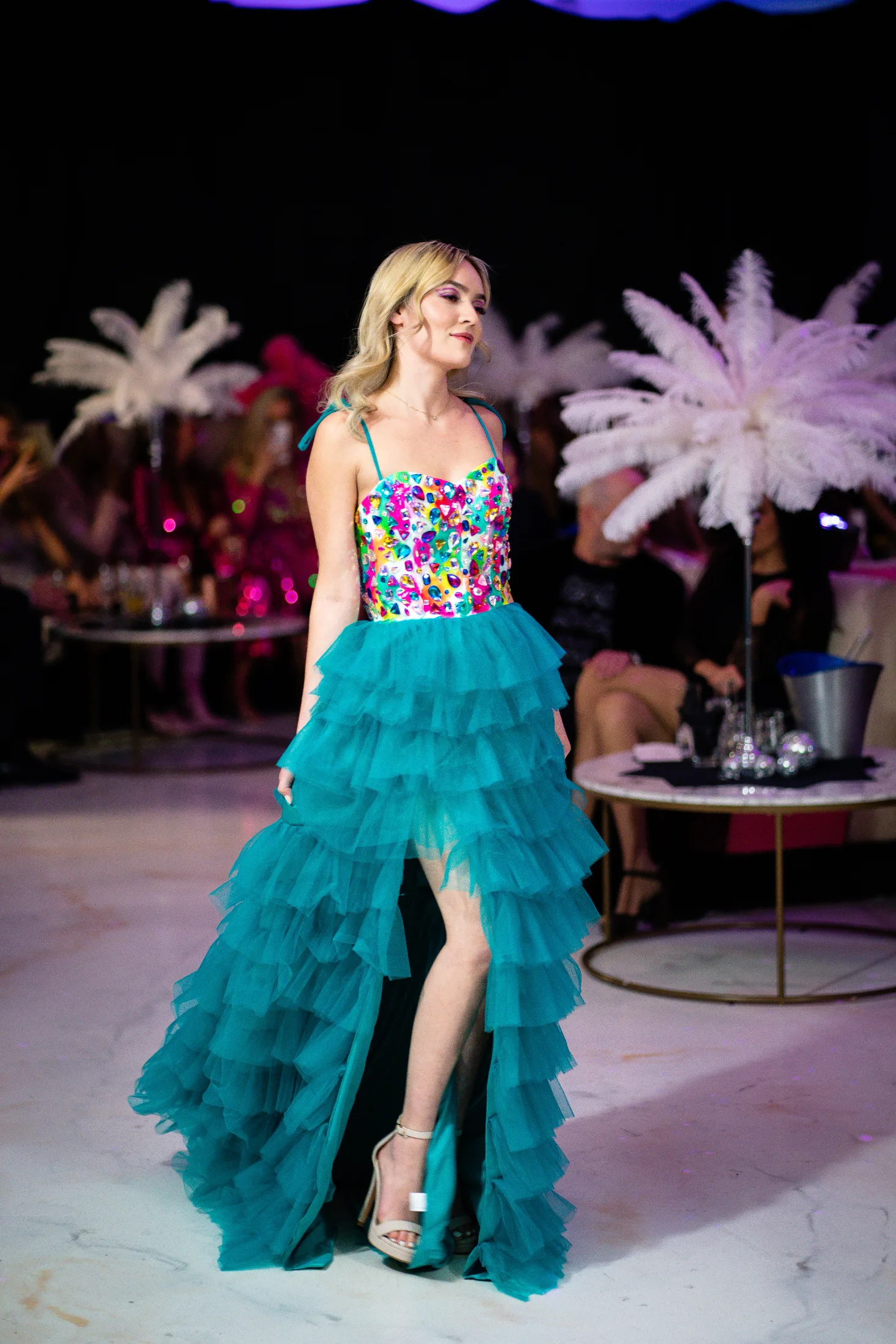 A model in a jewelled bodice and tiered teal tulle gown walks past guests at a lounge-style show