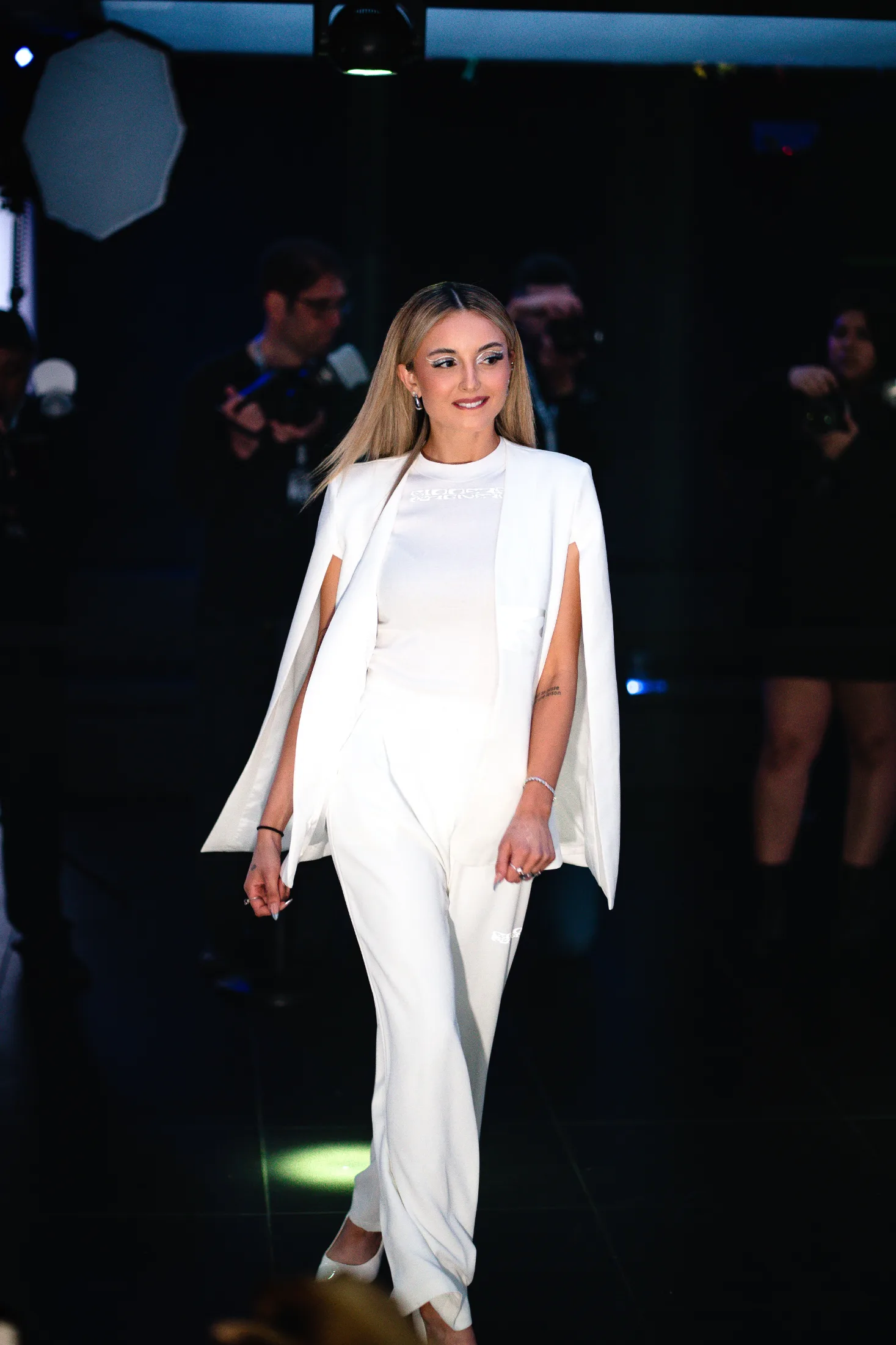 A model in a white cape blazer and wide trousers walks past photographers